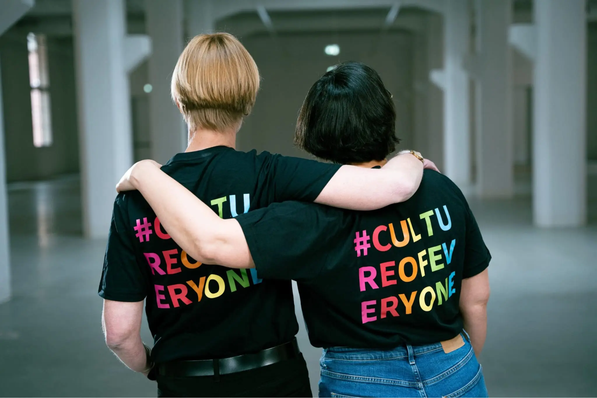 Two people seen from behind, holding each other arm in arm and wearing black T-shirts with the colorful or rainbow-colored inscription “Culture of Everyone”
